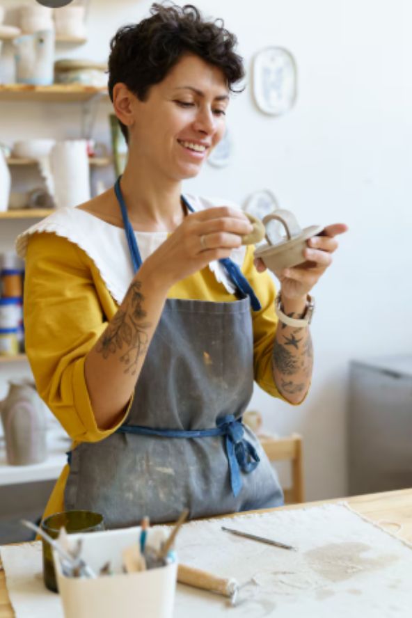 Uma mulher sorridente com tatuagens no braço e avental cinza manipula uma pequena peça de argila clara em um ateliê iluminado, com prateleiras de cerâmicas ao fundo.