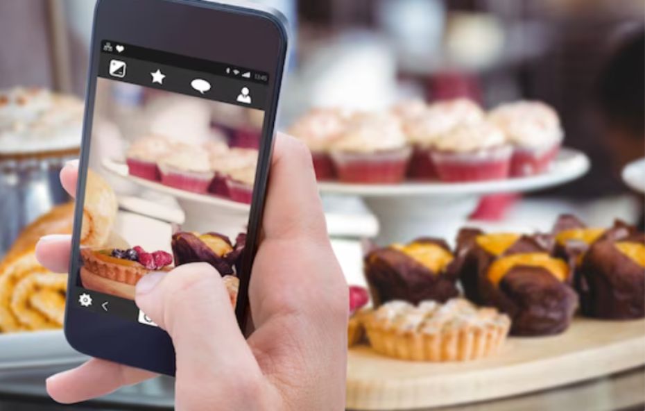Uma mão segura um smartphone em primeiro plano para fotografar uma vitrine repleta de doces variados como cupcakes e tortas em uma cafeteria moderna.