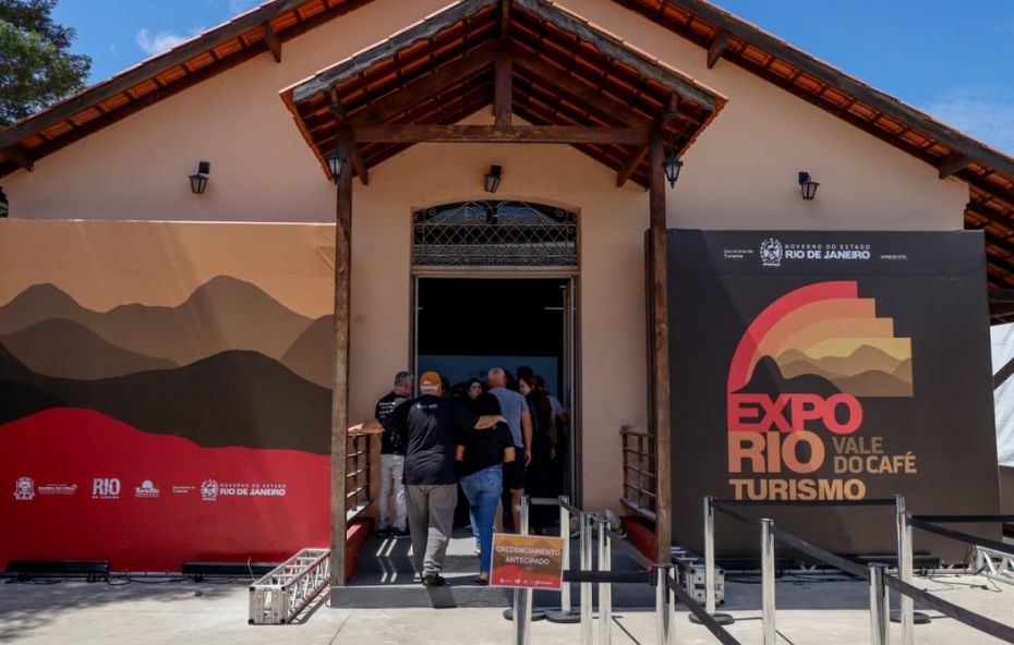 Pessoas entram em um casarão histórico restaurado que serve como entrada para o evento ExpoRio Vale do Café Turismo com painéis promocionais nas cores vermelho preto e laranja.