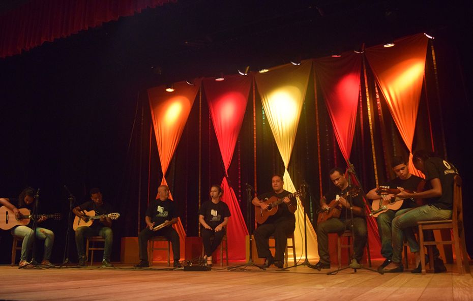 Fotografia de um grupo de jovens e adultos sentados em um palco tocando violão e percussão durante uma apresentação cultural com iluminação cênica colorida ao fundo.