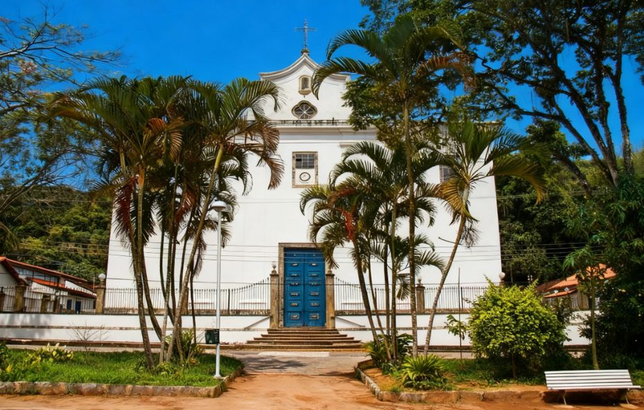 Fachada de uma igreja colonial branca com porta azul central e palmeiras à frente sob um céu azul limpo em Conservatória.