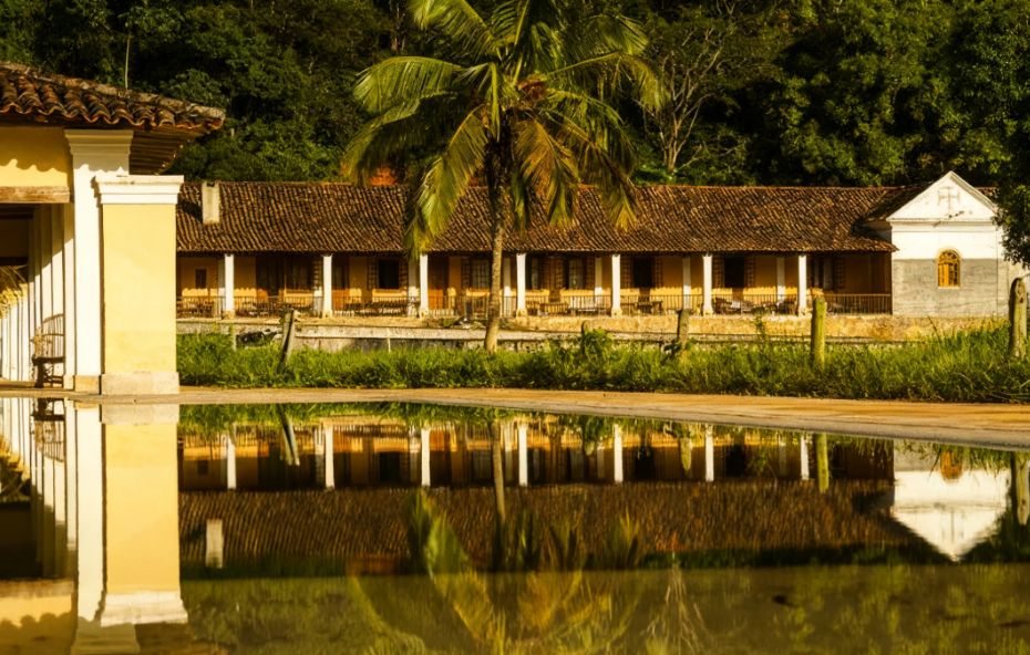 Fotografia da Fazenda Alliança mostrando um longo casarão amarelo e branco com colunatas e telhado de barro refletido em um espelho d'água em primeiro plano sob luz solar quente.