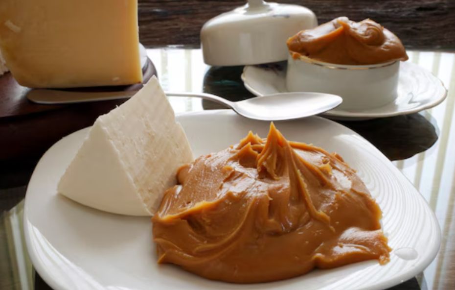 Close de um prato branco com uma porção generosa de doce de leite pastoso e uma fatia de queijo branco ao lado. Ao fundo há uma peça maior de queijo curado e um pequeno recipiente de porcelana também servido com o doce sobre uma mesa de vidro.