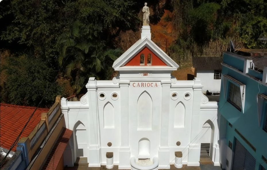 Fotografia frontal de um chafariz histórico branco com detalhes em vermelho e uma estátua no topo localizado entre prédios em Barra do Piraí com a inscrição Carioca no centro.
