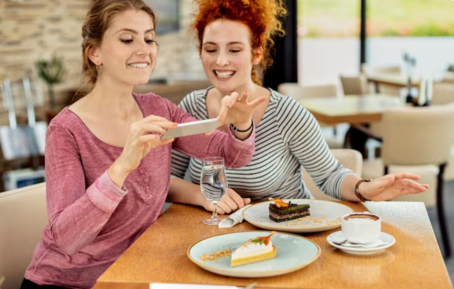 Duas amigas sorridentes sentadas à mesa de um restaurante moderno enquanto uma delas usa o celular para fotografar fatias de tortas gourmet e uma xícara de café.