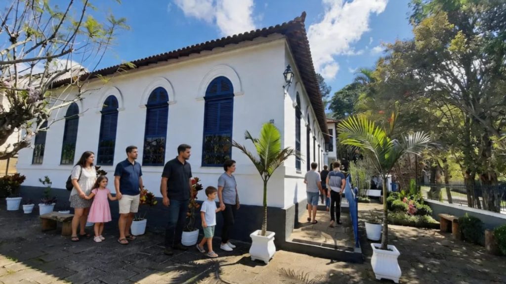 Uma fotografia colorida da fachada de uma casa colonial preservada, identificada como a entrada do Restaurante Adolphina. A construção de dois andares tem paredes brancas caiadas, janelas com molduras e persianas azul-escuras e um telhado de terracota. No primeiro plano, uma família de seis pessoas, incluindo adultos e crianças, caminha em um caminho de pedras em direção à entrada principal. Vasos de plantas brancos e bancos de madeira rústicos decoram a área. Ao fundo, um grupo de pessoas sobe uma rampa azul em direção a uma entrada lateral. O ambiente é cercado por vegetação exuberante e árvores frondosas sob um céu azul claro com nuvens.