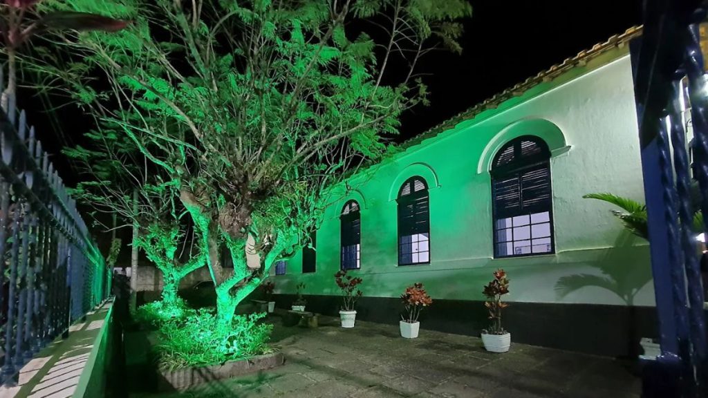 Fotografia noturna da fachada lateral do Restaurante Adolphina em Ipiabas. A parede branca da construção histórica está iluminada por luzes verdes que também atingem as copas das árvores à frente. As janelas coloniais possuem molduras arqueadas e persianas escuras. No pátio de pedras, há pequenos vasos de plantas brancos alinhados. À esquerda, vê-se uma grade de ferro escura que delimita a propriedade.