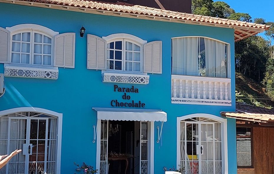Fotografia da fachada de um casarão de dois andares pintado em azul vibrante com detalhes em branco. O estabelecimento possui janelas de madeira com venezianas abertas e uma placa central com o nome Parada do Chocolate acima da porta de entrada que conta com um toldo branco.