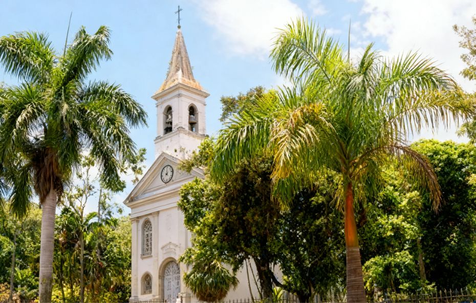 Fotografia da fachada branca em estilo neoclássico da Catedral de Nossa Senhora de Sant'Ana em Barra do Piraí cercada por palmeiras e árvores sob um céu claro com nuvens.