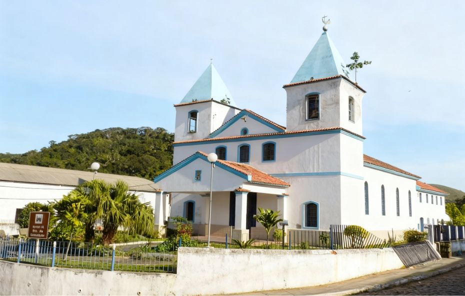 Vista frontal da Igreja de Nossa Senhora da Conceição com paredes brancas detalhes em azul duas torres com telhados pontiagudos e um pequeno jardim cercado à frente sob céu claro.