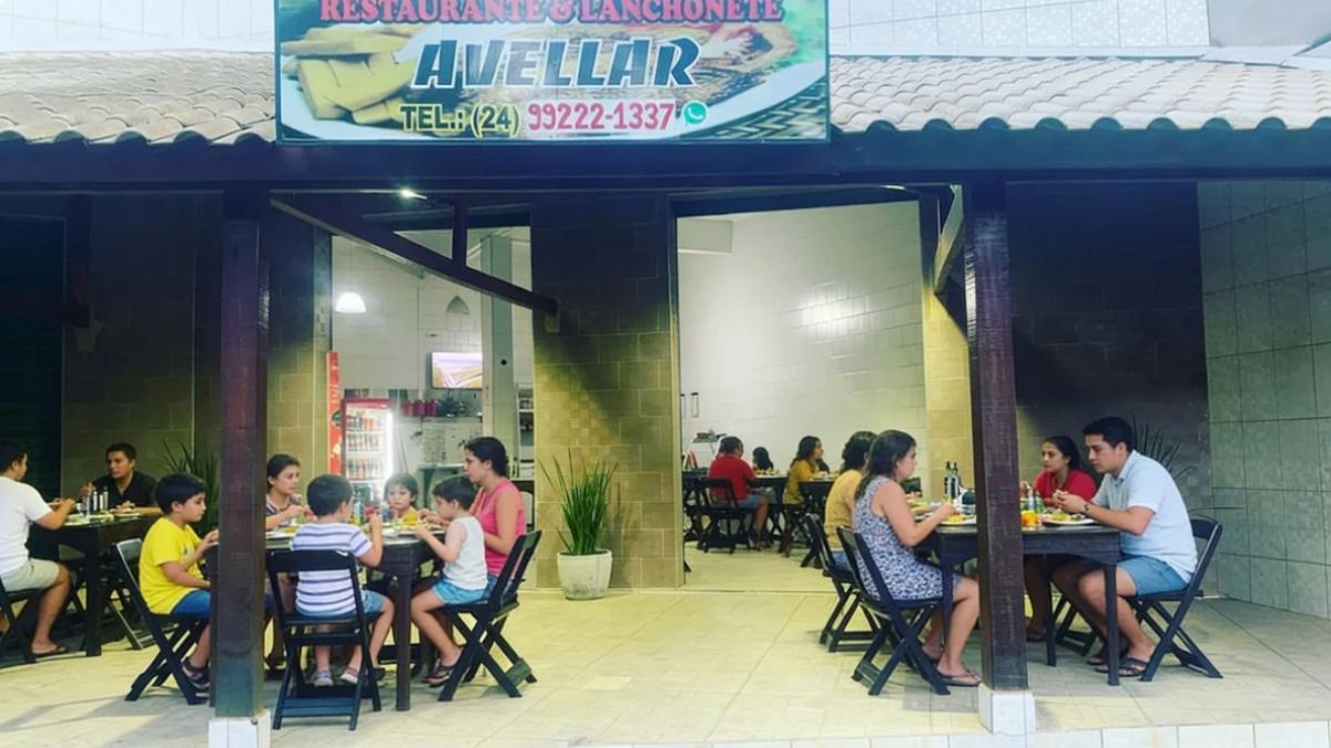 Fotografia colorida da fachada de um restaurante chamado Avellar durante o período noturno. O estabelecimento possui uma varanda coberta com telhas de barro e vigas de madeira escura. Várias mesas de madeira preta estão ocupadas por famílias e casais jantando. No topo, há uma placa com o nome do restaurante e um número de telefone com o código de área vinte e quatro. O piso é de cerâmica clara e as paredes externas são revestidas com blocos cinzas. A iluminação interna é branca e brilhante.
