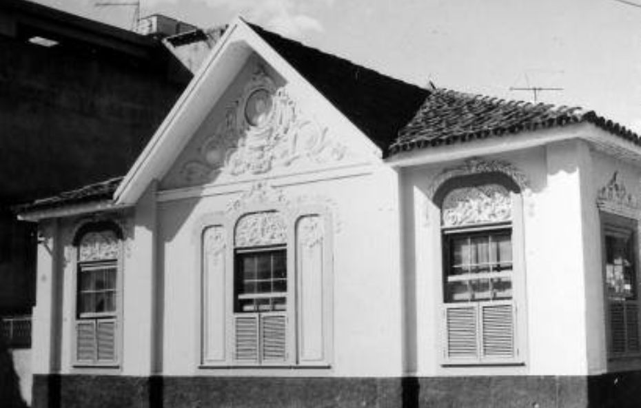 Fotografia antiga em preto e branco de uma pequena construção religiosa com ornamentos detalhados no frontão triangular e janelas de madeira trabalhadas.