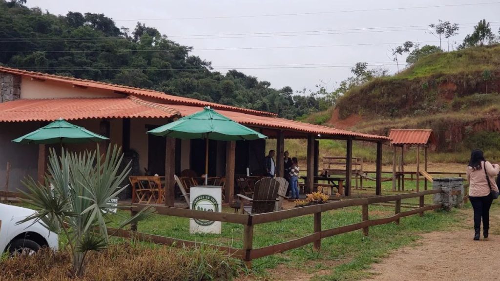 Fotografia da área externa de um restaurante de campo com telhado de barro e varanda de madeira. Há guarda-sóis verdes e uma placa indicando frango com quiabo e costela aos sábados e feijoada aos domingos. O ambiente é cercado por gramado e montanhas.
