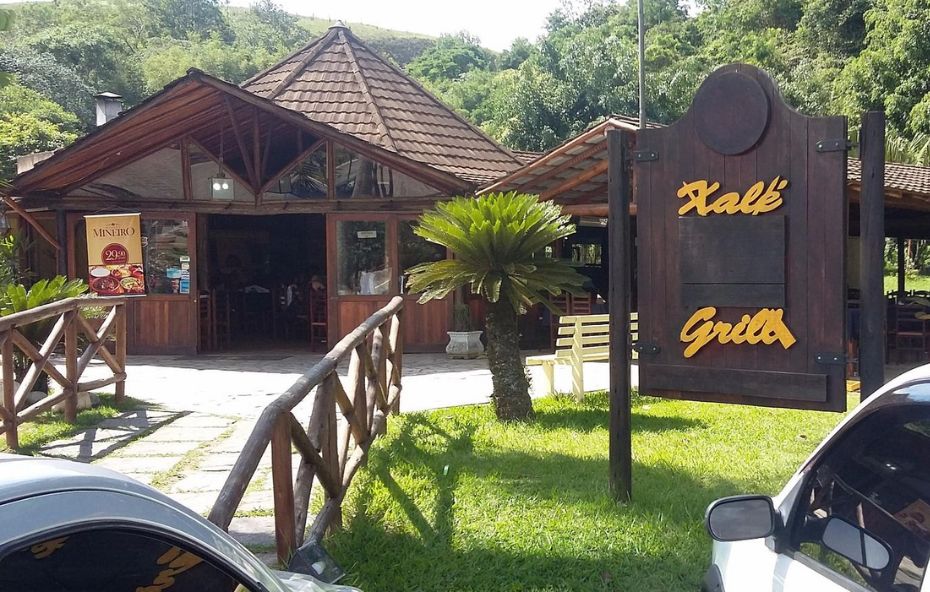 Fachada de um restaurante de madeira com telhado em formato piramidal cercado por jardins e montanhas. Uma placa de madeira escura com letras amarelas indica o nome Xalé Grill na frente de um gramado ensolarado.