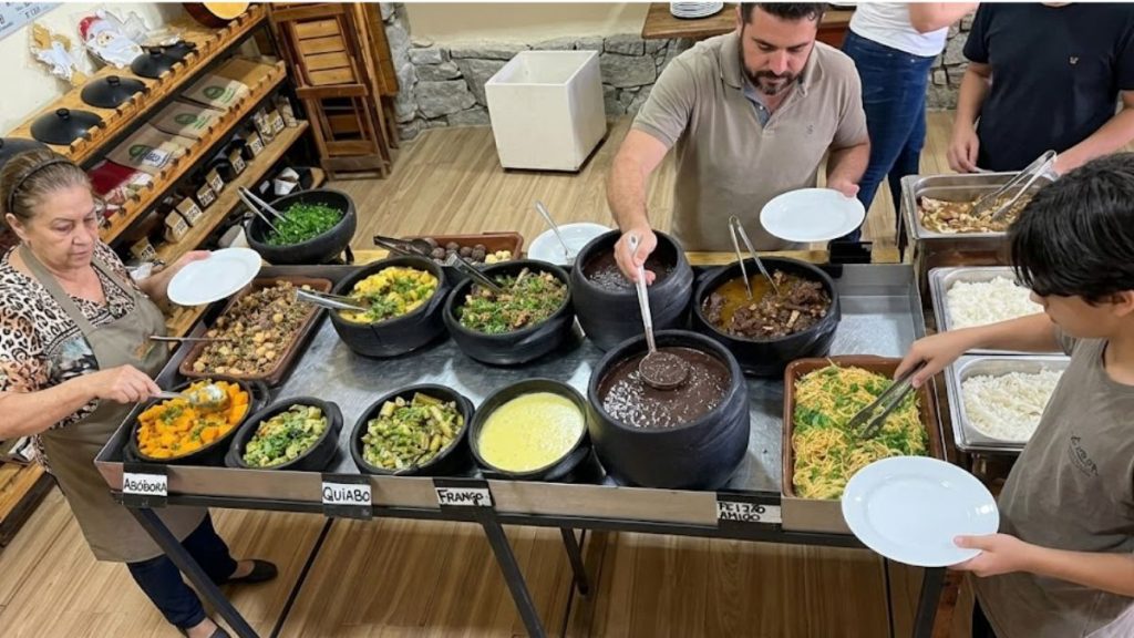 Pessoas se servem em um buffet de comida mineira com diversas opções dispostas em panelas de barro sobre um balcão térmico. Há pratos como feijoada, frango com quiabo, abóbora e arroz. O ambiente possui chão de madeira e prateleiras rústicas ao fundo.