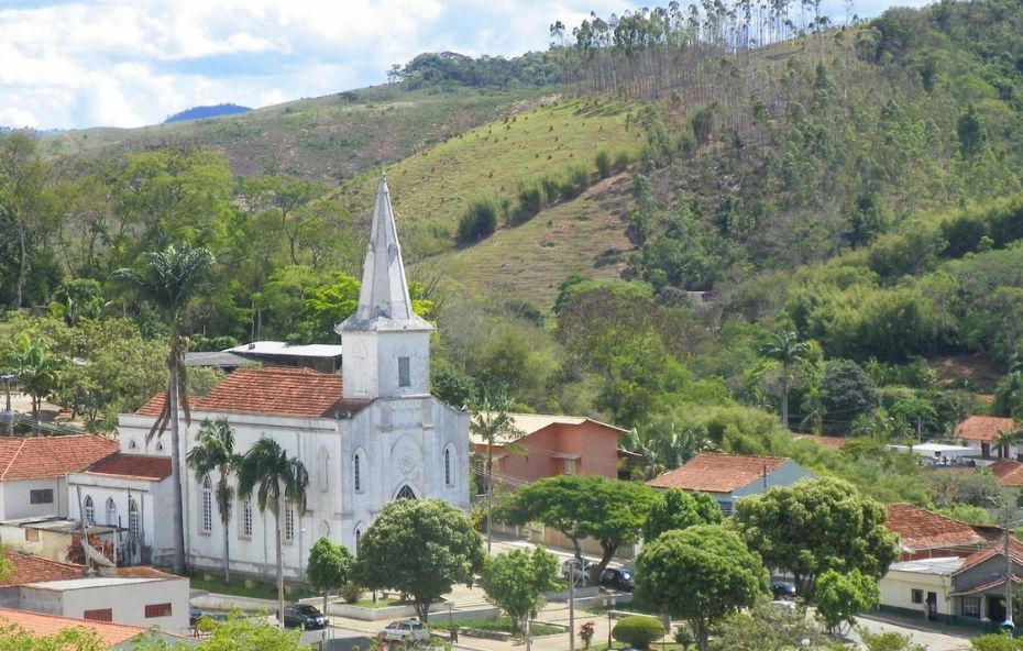 Vista aérea de uma igreja branca de estilo neogótico com torre pontiaguda cercada por casas de telhados cerâmicos e colinas verdes em Rio das Flores.