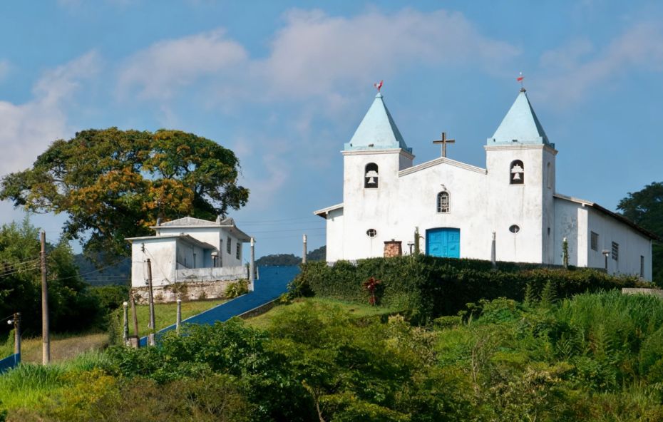 Vista da Igreja Nossa Senhora da Soledade com suas duas torres brancas de telhado azul localizada em um plano elevado cercada por vegetação verde sob céu claro.