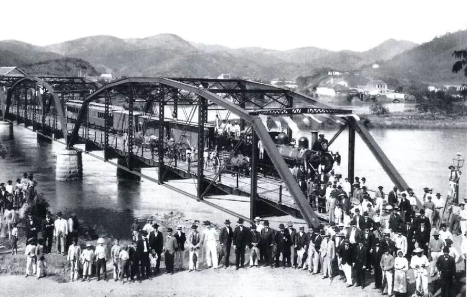 Fotografia antiga em preto e branco mostrando uma multidão de pessoas vestidas formalmente em frente a uma ponte metálica com uma locomotiva a vapor e vagões parados sobre ela.
