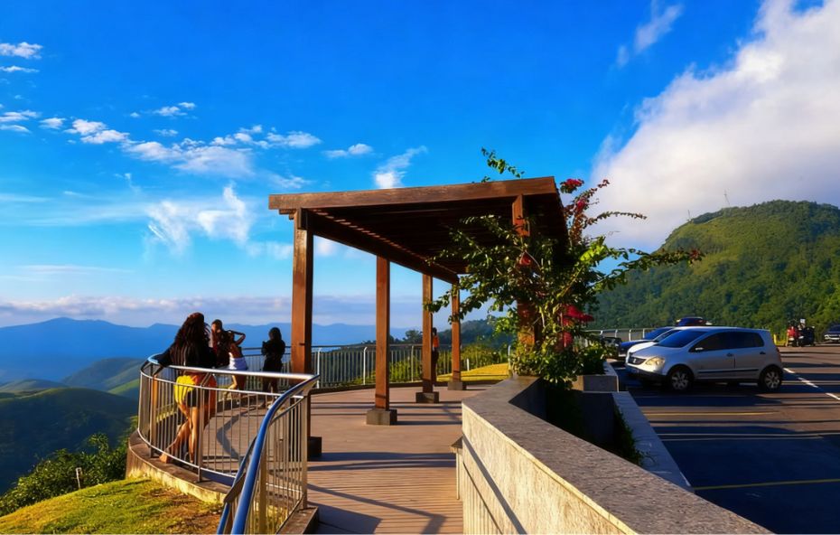 Pessoas em um mirante com deck de madeira e guarda-corpo de metal observando montanhas verdes sob um céu azul com nuvens em Conservatória.