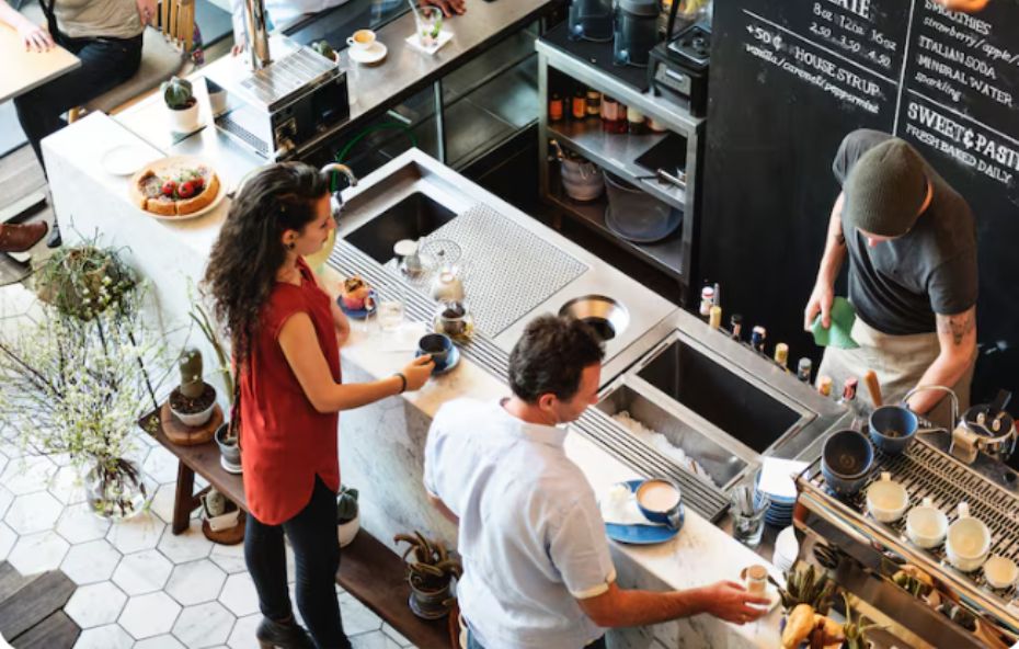 Vista aérea de um balcão de cafeteria moderno em mármore branco com clientes sendo atendidos por um barista enquanto aguardam seus cafés e doces.