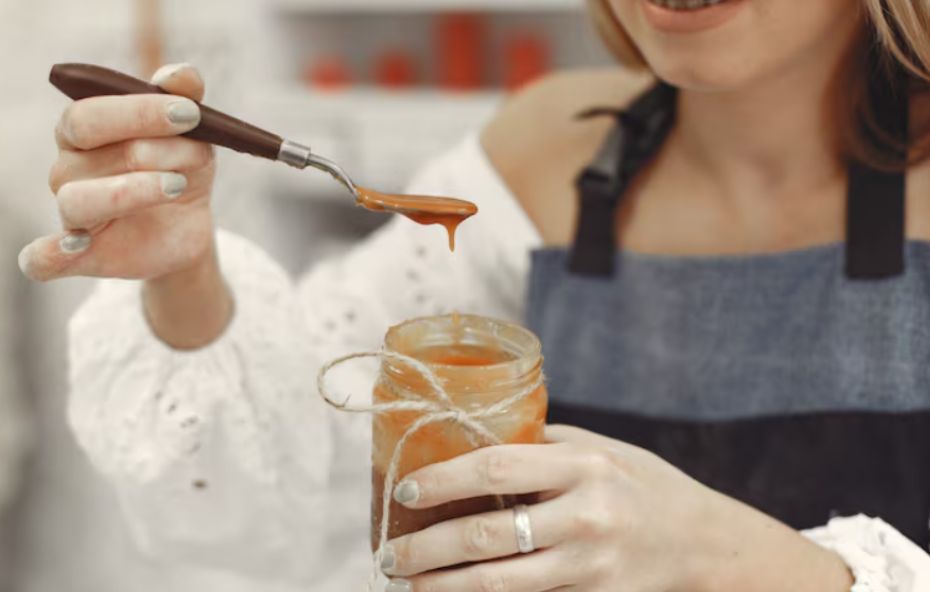 Close das mãos de uma pessoa segurando um pote de vidro com doce cremoso decorado com um laço de corda. Com a outra mão a pessoa levanta uma pequena espátula de onde escorre uma gota de calda caramelizada. A pessoa veste um avental jeans e uma blusa branca.