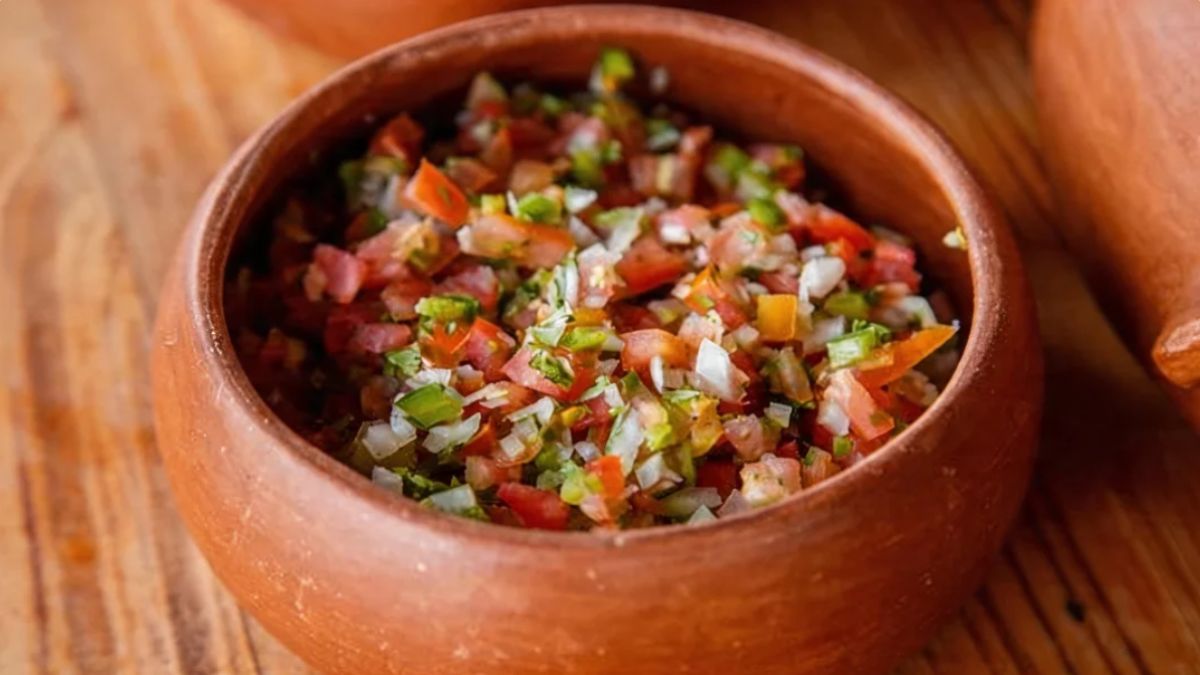 Uma tigela redonda de barro cozido repleta de vinagrete picadinho com tomates cebolas e cheiro verde sobre uma mesa de madeira.