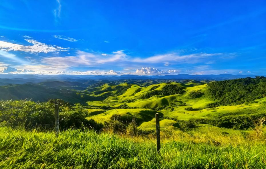 Vista panorâmica de montanhas verdes onduladas sob um céu azul vibrante com nuvens brancas em Conservatória.