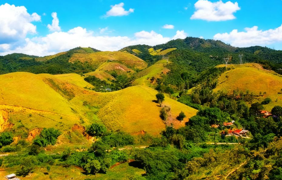 Vista panorâmica de colinas verdes e montanhas da Serra do Mar sob céu azul com nuvens brancas e pequenas casas integradas à vegetação em Engº Paulo de Frontin.
