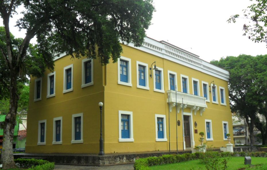 Edifício histórico de dois andares pintado em amarelo vibrante com janelas de molduras brancas e detalhes em azul cercado por jardins e árvores em Piraí.