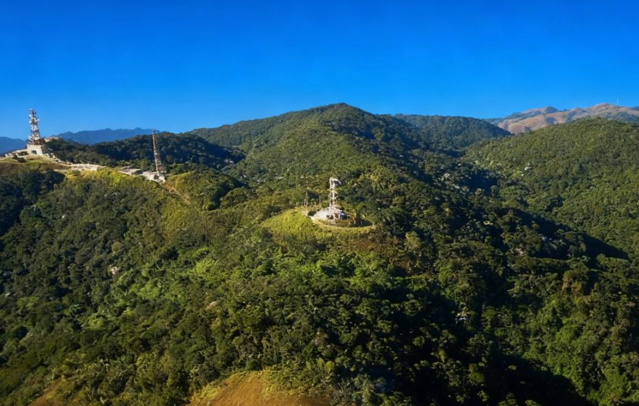 Vista aérea de montanhas cobertas por vegetação densa de Mata Atlântica sob céu azul com torres de comunicação instaladas nos cumes em Engº Paulo de Frontin.