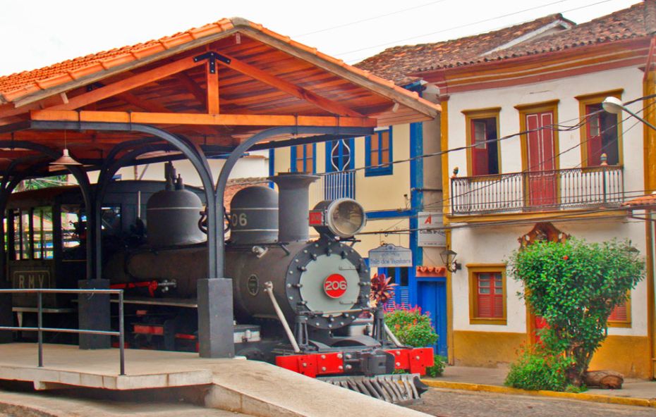 Locomotiva a vapor antiga em exposição sob uma plataforma de telhado de madeira ao lado de casarões coloniais coloridos em Conservatória.