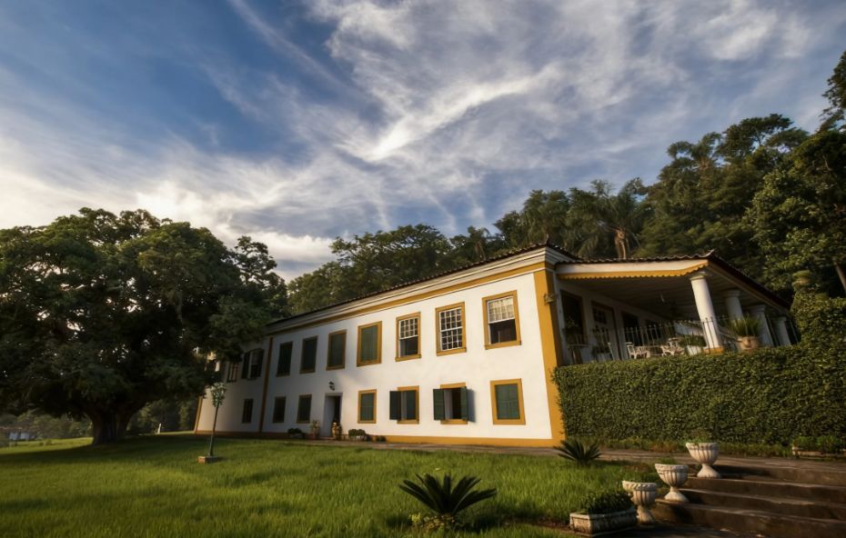 Casarão colonial de dois andares com fachada branca e janelas amarelas cercado por gramado verde e árvores sob um céu com nuvens em Valença.