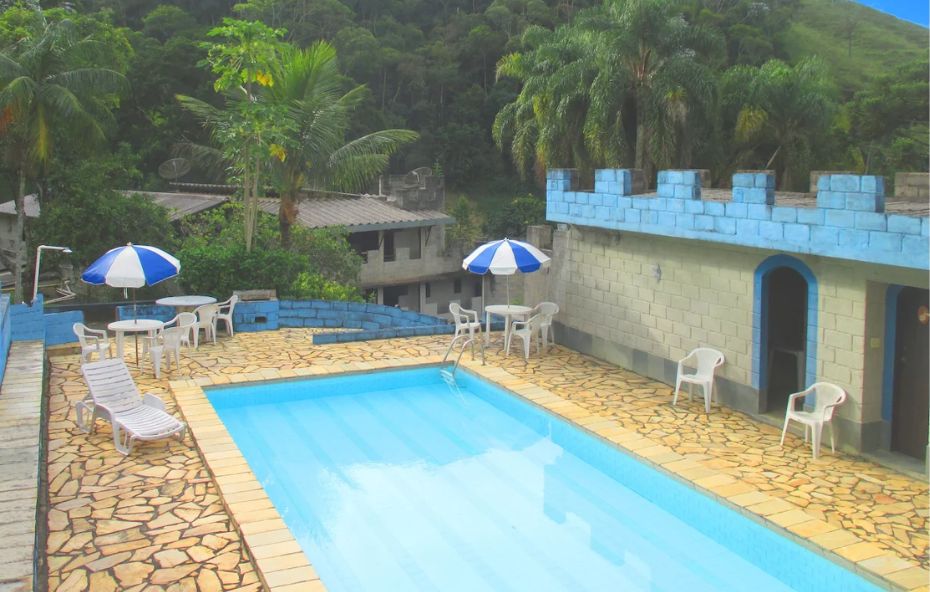 Piscina ao ar livre no castelo de Ipiabas com área de lazer, cadeiras, mesas com guarda sol e vegetação da serra ao fundo.