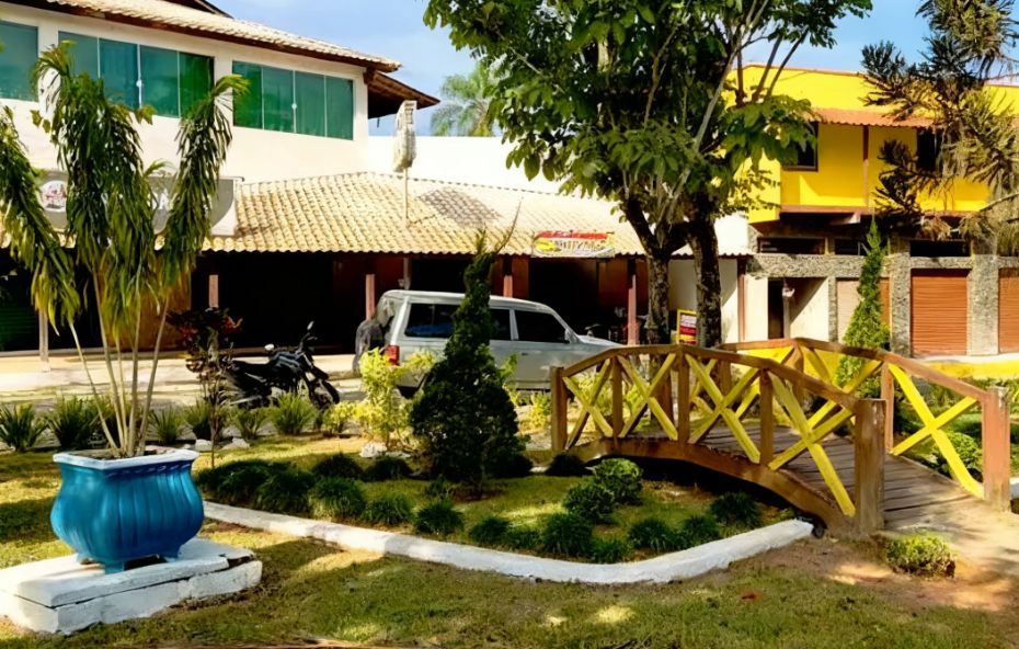 Área externa em Ipiabas com uma pequena ponte de madeira ornamental amarela sobre um jardim gramado em frente a construções comerciais e pousadas.