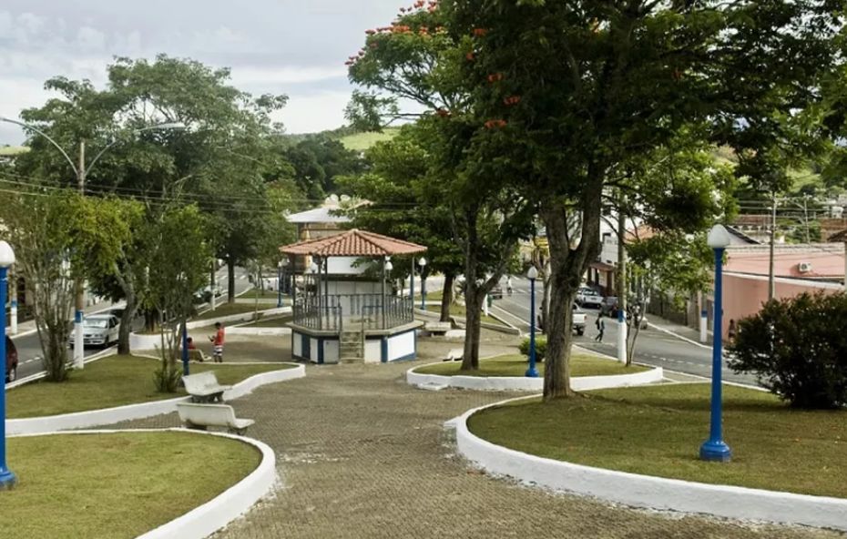 Vista aérea de uma praça arborizada com coreto central de telhado colonial, canteiros circulares gramados, bancos brancos e postes de iluminação azuis em Rio Claro.