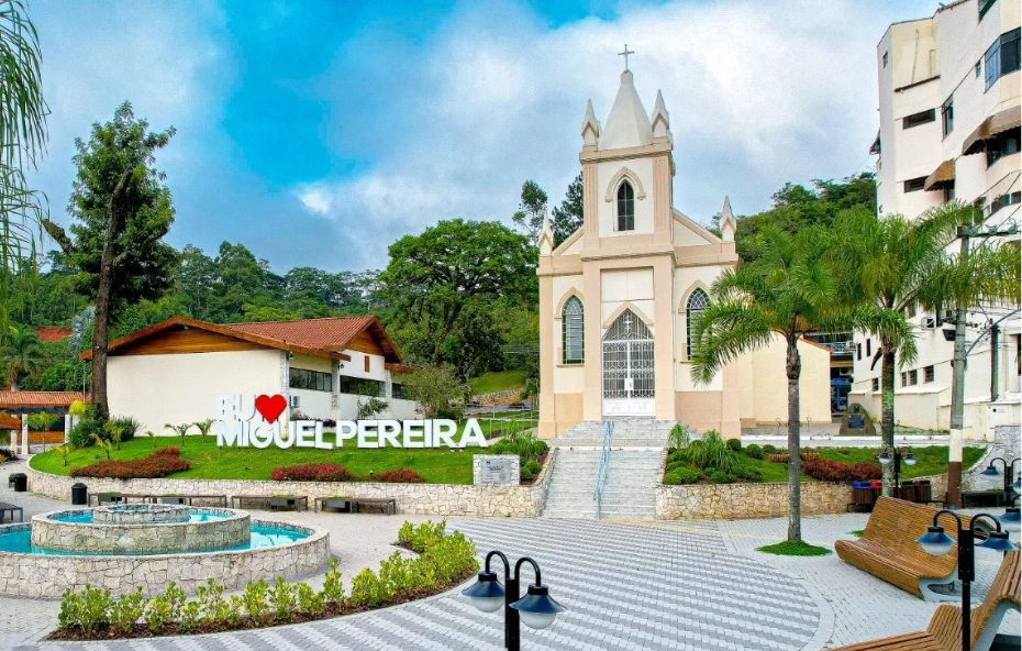 Praça pavimentada com uma fonte circular à esquerda, letreiro com a frase Eu amo Miguel Pereira ao centro e uma igreja de estilo neogótico ao fundo sob céu nublado.