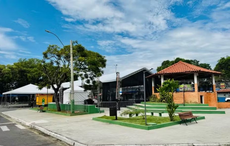 Praça urbana com quiosque de telhado colonial, canteiros verdes, bancos de madeira e estruturas de tendas brancas para eventos sob um céu com nuvens.