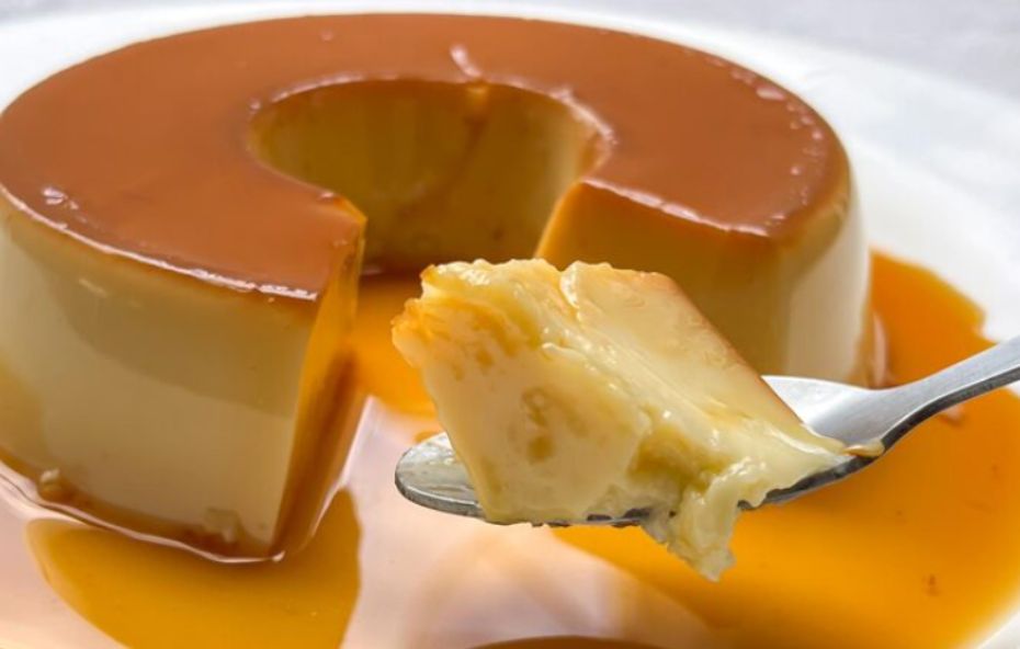 Visão aproximada de um pudim de leite condensado cremoso com calda de caramelo brilhante e uma colher retirando um pedaço suculento.