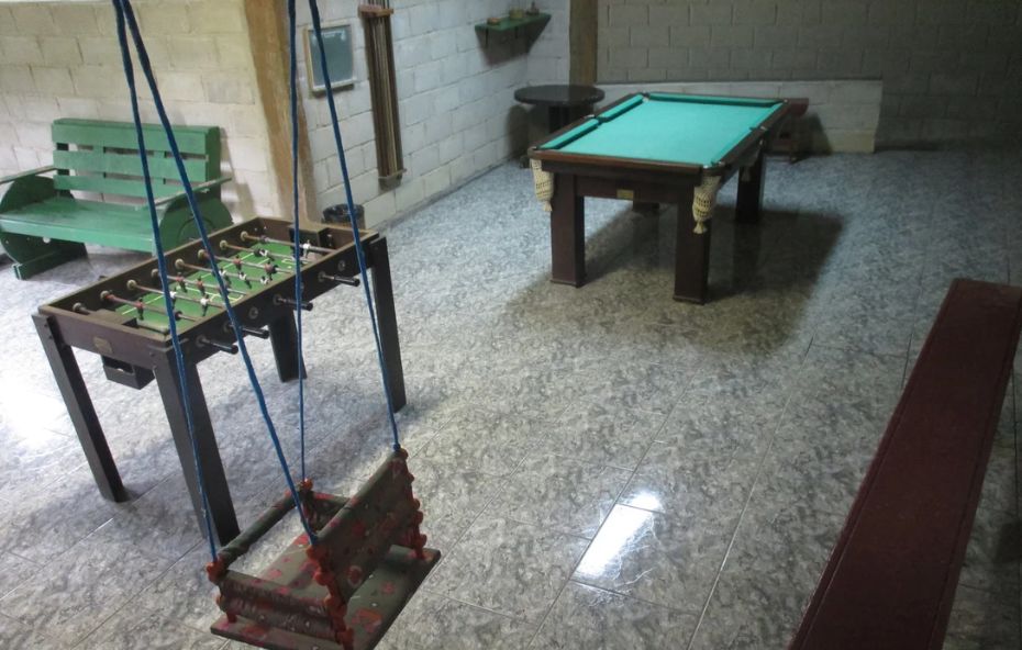 Sala de jogos com mesa de sinuca, mesa de pebolim, balanço artesanal e bancos em ambiente fechado com piso de cerâmica.