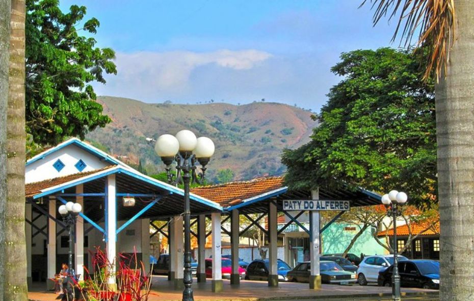 Vista de uma estação com coberturas metálicas azuis e brancas onde se lê o nome Paty do Alferes cercada por palmeiras e montanhas ao fundo sob céu azul.