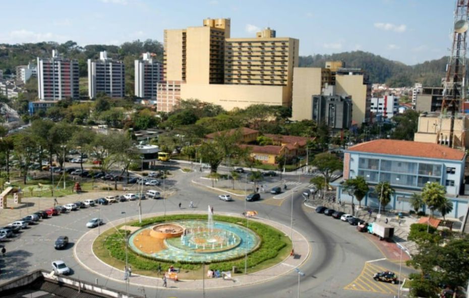 Fotografia aérea de uma grande rotatória com fonte luminosa central cercada por prédios altos e ruas movimentadas com carros estacionados em Volta Redonda sob céu claro.