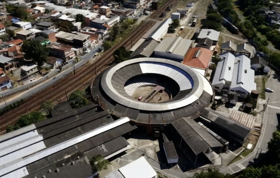 Fotografia aérea de uma grande estrutura circular de manutenção ferroviária com teto metálico cinza e branco cercada por trilhos e galpões industriais no centro urbano de Barra do Piraí.