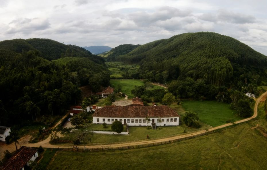 Fotografia aérea de um casarão colonial branco e comprido com telhado de barro cercado por vastos campos verdes e montanhas cobertas por florestas em Barra do Piraí.