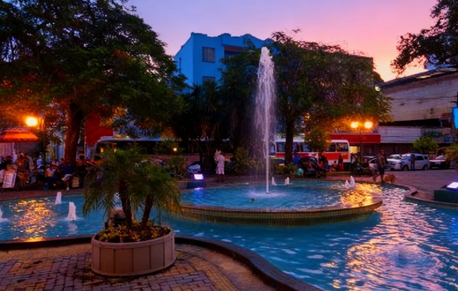 Fotografia ao entardecer da Praça Nilo Peçanha em Barra do Piraí mostrando um chafariz circular central com jatos de água iluminados e árvores ao redor sob um céu em tons de rosa e roxo.