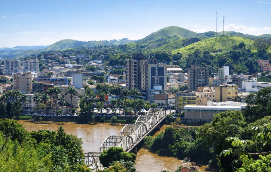 Fotografia aérea de Barra do Piraí mostrando o Rio Paraíba do Sul atravessado por uma ponte metálica cinza, cercado por edifícios e morros verdes.