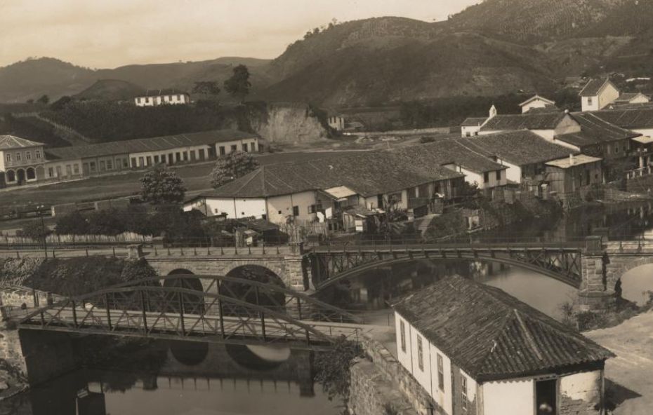 Fotografia antiga em tons de sépia mostrando um conjunto de pontes de pedra e ferro sobre um rio com casarões coloniais e morros ao fundo.