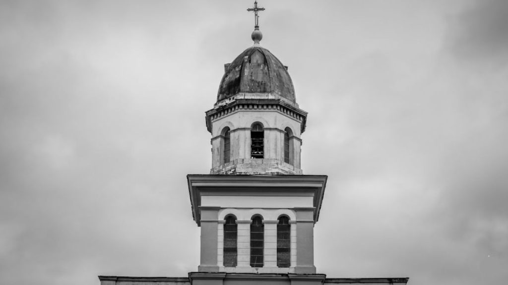 Fotografia em preto e branco centralizada na parte superior da torre da Igreja Nossa Senhora da Piedade em Ipiabas. A imagem destaca três janelas estreitas em arco no corpo inferior da torre e três aberturas superiores que revelam o sino. O topo é composto por uma cúpula arredondada com uma cruz metálica sobreposta a um céu nublado que preenche todo o fundo.