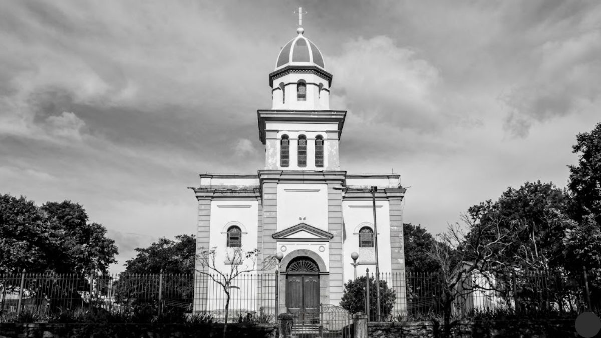 Fotografia em preto e branco com ângulo de visão centralizado da Igreja Matriz de Nossa Senhora da Piedade, em Ipiabas, Barra do Piraí. A imagem captura a fachada simétrica da igreja, que tem uma torre central com cúpula e cruz. Há um muro de pedra e uma cerca de metal na frente da igreja. Árvores cercam o edifício e o céu está coberto de nuvens.