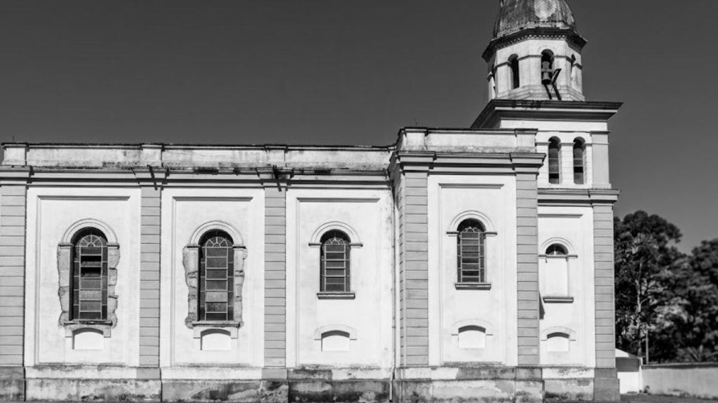 Uma fotografia em preto e branco com perspectiva lateral e centralizada da Igreja Matriz de Nossa Senhora da Piedade, em Ipiabas, Barra do Piraí. A imagem foca nos detalhes arquitetônicos e em quatro janelas laterais, sendo três de tamanho médio em arco e uma menor na torre. A torre central da igreja, com sua cúpula e cruz no topo, é visível à direita. O edifício é feito de alvenaria com detalhes em pedra e cal e está situado em um gramado sob um céu limpo. Uma cerca de metal e árvores cercam a propriedade.