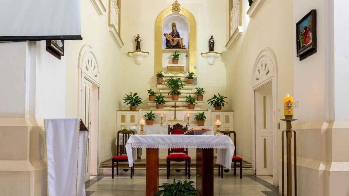 Fotografia colorida do interior da Igreja Nossa Senhora da Piedade em Ipiabas mostrando o altar principal centralizado. O altar possui uma mesa coberta com toalha branca rendada e vasos de plantas ornamentais ao fundo. Uma imagem sacra de Nossa Senhora da Piedade está em um nicho dourado no topo. Paredes laterais brancas com portas em arco e detalhes dourados completam a cena sob uma iluminação quente e acolhedora.